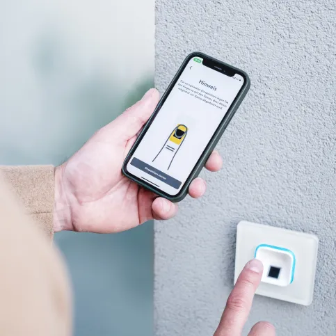 Mit der ekey bionyx App wird das ekey System verwaltet. Hand, die ein Smartphone hält, während sie einen modernen Türöffner mit Touchscreen und minimalistischer Gestaltung aktiviert.