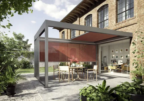 Terrasse mit freistehender Pergola aus Aluminium und Glas, umgeben von Pflanzen und historischem Backsteinbau.